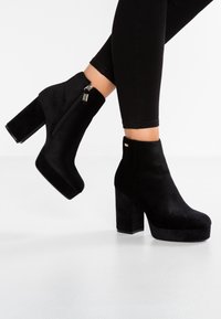 Bottines en daim noir avec un talon bloc haut, une plateforme avant et une fermeture éclair latérale. Texture lisse et design minimaliste.