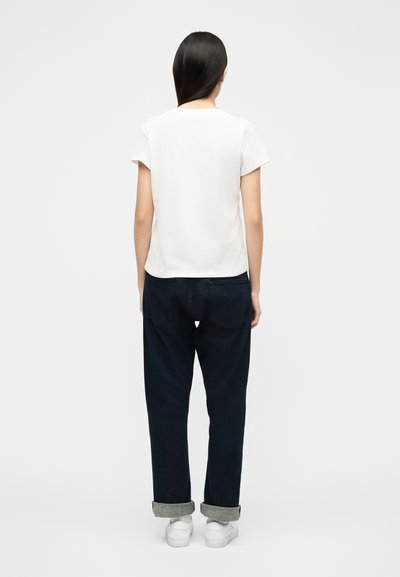 Γυναίκα με μακριά σκουρόχρωμα μαλλιά που φοράει απλό λευκό μπλουζάκι, σκούρο κατεβατό τζιν και λευκά αθλητικά παπούτσια, στέκεται πλάτη προς τα εμπρός σε λευκό φόντο.