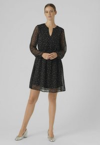 Robe noire à pois avec de longues manches transparentes, décolleté en V et taille froncée. Longueur au genou avec une jupe fluide et une texture de tissu légère.