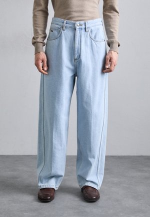 Persoon draagt lichtblauwe wijde spijkerbroek, bruine leren schoenen en een beige top met lange mouwen, staand op een grijze vloer.