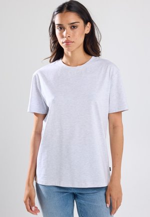 Jonge vrouw met donker haar, gekleed in een effen lichtgrijs T-shirt met korte mouwen en een blauwe spijkerbroek, tegen een effen achtergrond.