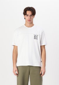 Hvit t-skjorte med korte ermer, rund hals og grafisk trykk som inkluderer "GUESS JEANS 81" på venstre bryst. Har søm detaljer.