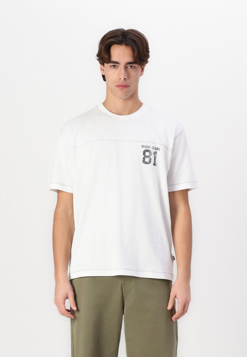 Hvit t-skjorte med korte ermer, rund hals og grafisk trykk som inkluderer "GUESS JEANS 81" på venstre bryst. Har søm detaljer.