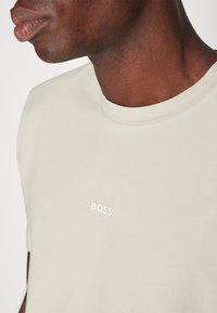 T-shirt beige clair en coton doux, avec un col rond et des manches courtes. Le nom de la marque "BOSS" est imprimé en blanc.