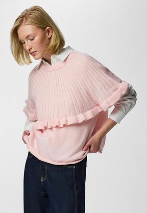 Femme blonde portant un poncho en tricot rose clair à volants par-dessus une chemise blanche boutonnée et un jean bleu foncé, debout les mains sur les hanches.