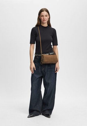 Kobieta ubrana w czarną bluzkę z krótkim rękawem, ciemnoniebieskie szerokie spodnie, czarne szpiczaste buty, pasek z ćwiekami i brązową torebkę na ramię, stojąca na jednolitym tle.