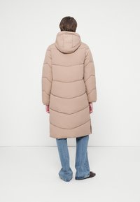 Persona de pie con la espalda visible, vistiendo un abrigo largo acolchado beige con capucha, jeans azules y zapatos oscuros, contra un fondo blanco plano.