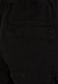 Svart denimtyg med en strukturerad yta. Har en bakficka med sömmar som stänger, vilket betonar en praktisk design och ett hållbart material.
