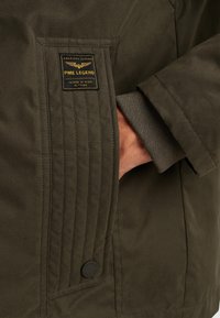 Olivově zelená bunda s texturovanou látkou, prošitými detaily, černým kulatým knoflíkem a prošitou nášivkou s logem vpředu.