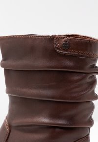 Brun läderstövel med en slouch-design, med stygnaccent och en metallknappdetalj vid vristen. Slät yta och medelhög.