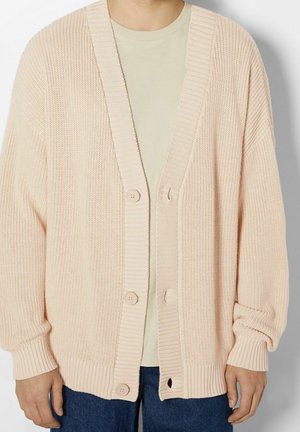 Cardigan lavorato a maglia di un rosa chiaro con scollatura a V, bordi a costine e tre grandi bottoni, indossato sopra una maglietta beige e jeans blu.