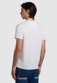 Maglietta bianca a maniche corte con colletto tondo, vista dal retro. Il tessuto appare liscio, caratterizzato da un design semplice e minimalista.