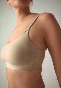 Vrouw draagt een beige naadloze beha met dunne bandjes, zijaanzicht tegen een effen lichte achtergrond.