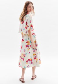 Robe imprimée florale en crème avec des manches courtes et une jupe plissée. Présente des fleurs rouges, roses et vertes vives. Portée avec des sandales marron.