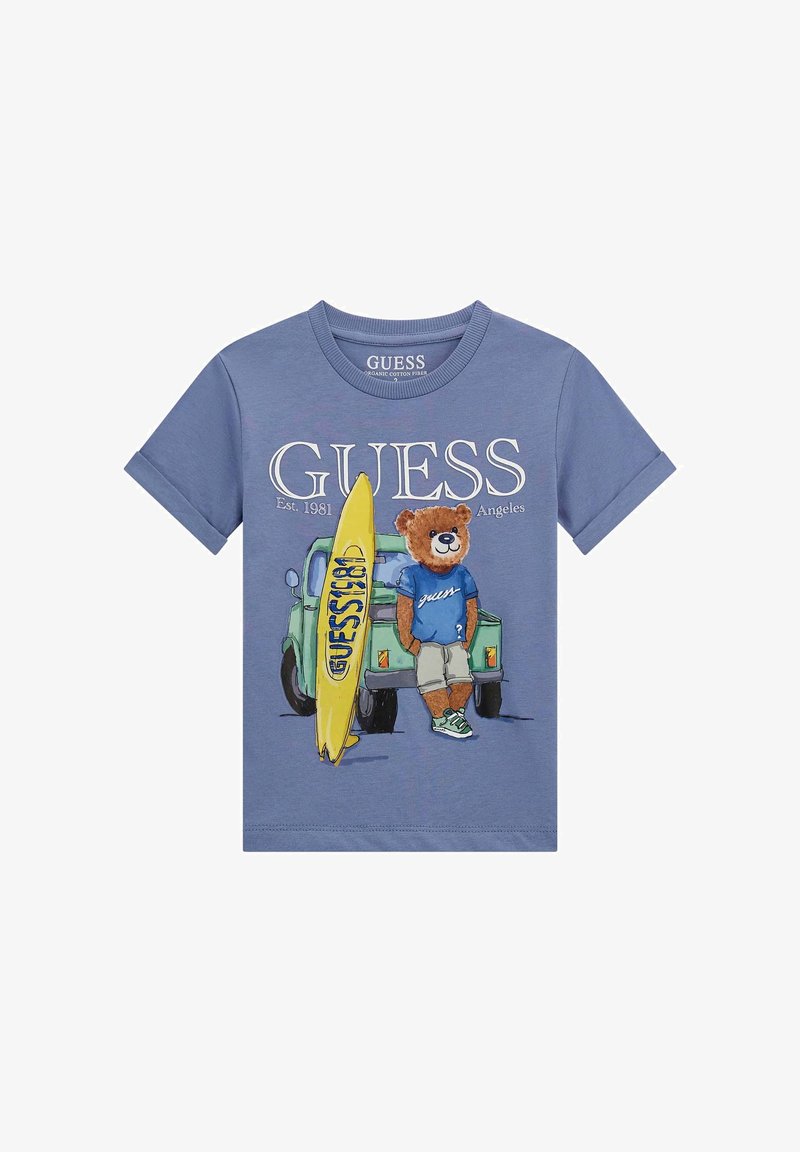Plava pamučna majica s grafikom medvjeda s daskom za surfanje, u kombinaciji s ležernim šorcem i tenisicama, te otisnutim "GUESS" logotipom iznad.