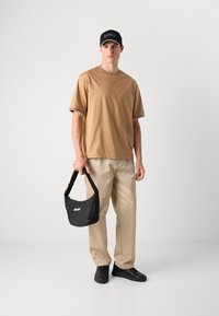 Ung mann står og har på seg en beige t-skjorte, beige bukser, svarte joggesko og caps, holder en svart BOSS skulderveske i stroppen med høyre hånd.