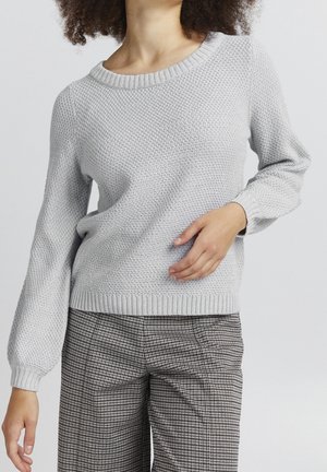 Femme portant un pull en maille texturé gris clair et un pantalon à carreaux noir et blanc, une main posée sur sa taille.