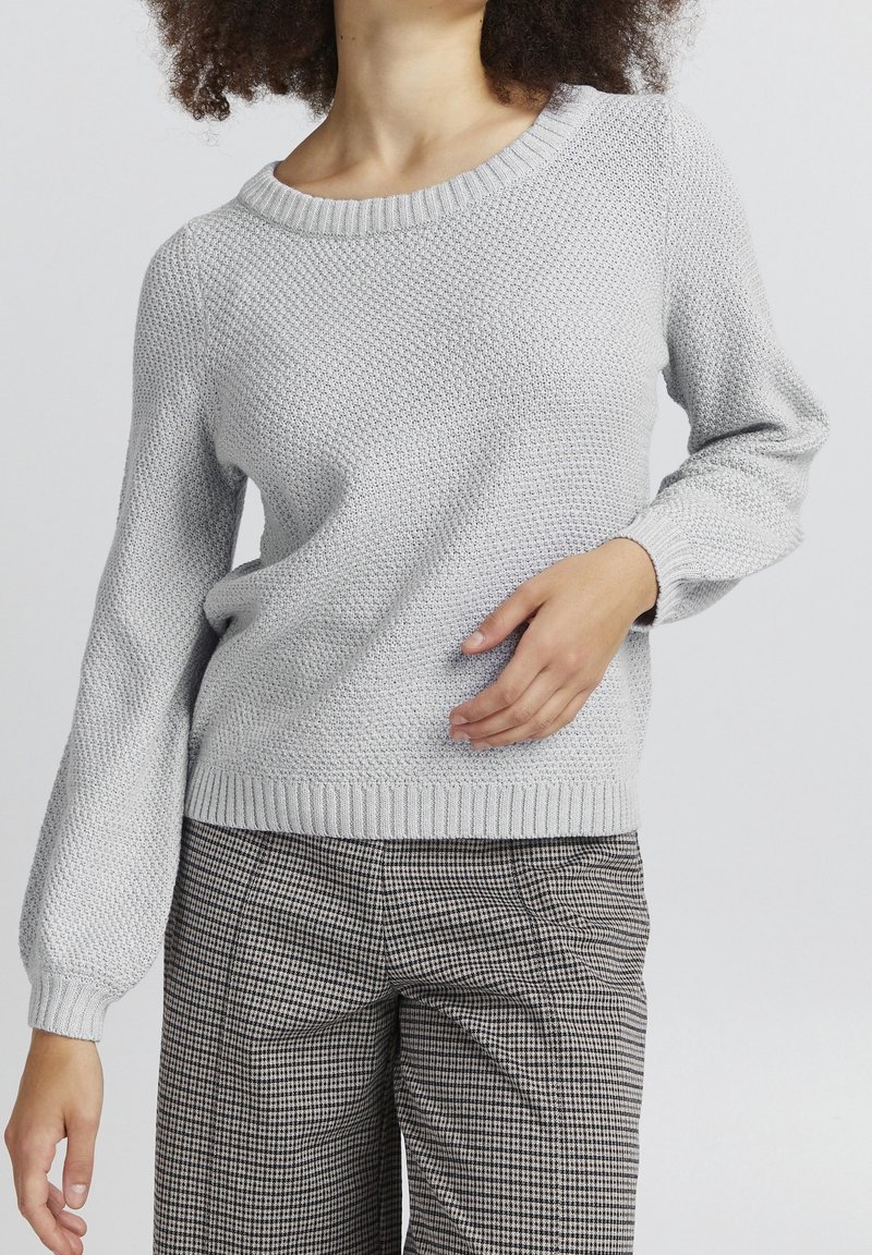 Femme portant un pull en maille texturé gris clair et un pantalon à carreaux noir et blanc, une main posée sur sa taille.