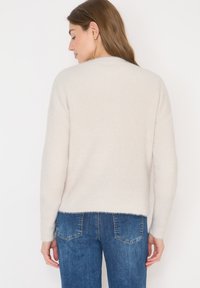 Helles beige Strickpullover mit gerippter Textur, langen Ärmeln und cropped Saum, kombiniert mit blauen Jeans.