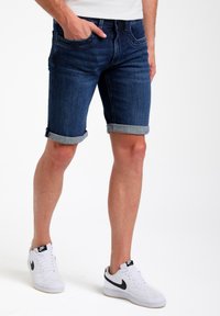 Pantalones cortos de mezclilla en azul oscuro con dobladillo vuelto. Combinados con zapatillas blancas con detalles en negro. El modelo se encuentra en una postura casual.