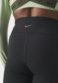 A fekete Nike leggings szoros illeszkedésű, magas derékú. A logó szürke színben jelenik meg, amit egy világoszöld felső egészít ki.