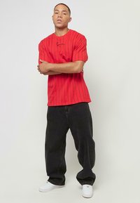 T-shirt rouge à manches courtes avec des rayures noires verticales et un petit logo brodé, associé à un jean noir ample et des baskets blanches.