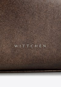 Bruin gestructureerd leren materiaal met een subtiele glans; beschikt over het gegraveerde logo "WITTCHEN" in zilverkleur.