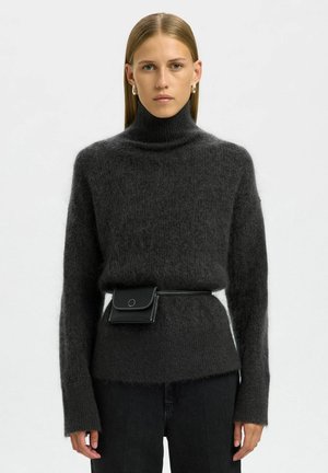 Donna con capelli lisci che indossa un maglione a collo alto grigio scuro e peloso, pantaloni neri, orecchini a cerchio e una borsa a cintura nera in vita.