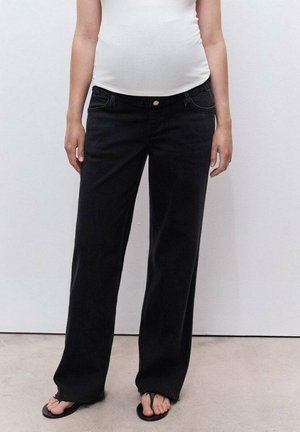 Person wearing a white top, black wide-leg jeans with raw hems, and black flip-flops standing against a plain light wall.