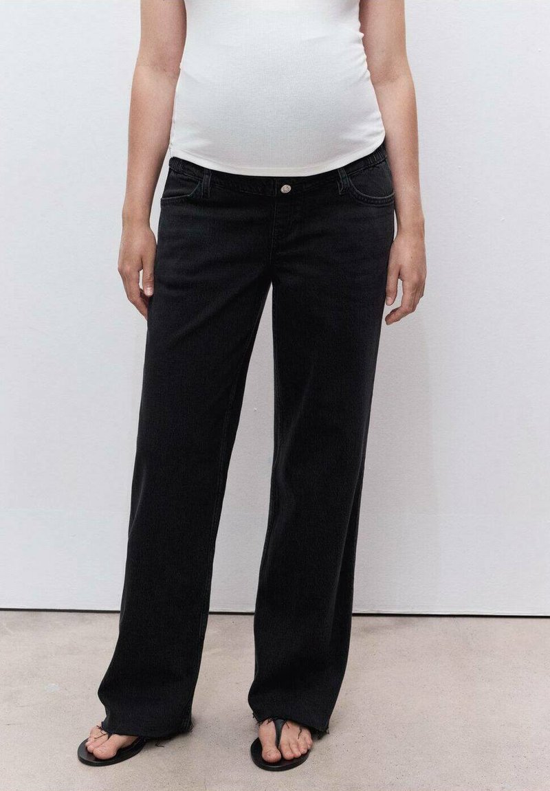 Persona vestida con una blusa blanca, jeans anchos negros con dobladillos deshilachados y chanclas negras, de pie contra una pared lisa clara.