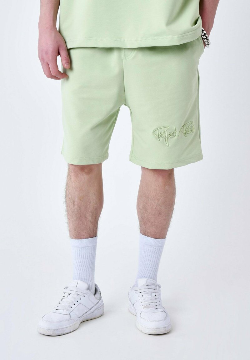 Shorts vert clair en tissu doux, avec un logo brodé sur le bas de la jambe gauche. Portés avec des chaussettes blanches et des baskets.