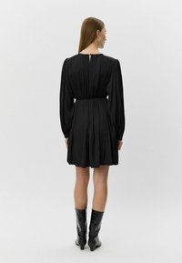 Juodas plisuotas suknelė su ilgomis rankovėmis, įtemptu liemeniu ir skylute nugaroje, derinama su juodomis krokodilų rašto batais iki blauzdų.