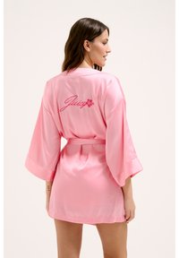 Robe en satin rose avec de larges manches et une ceinture à nouer, ornée de "Juicy" brodé en rose dans le dos, texture lisse et coupe mi-cuisse.