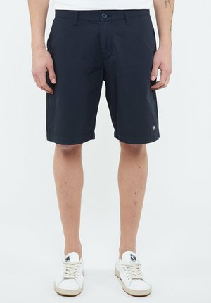 Hombre con pantalones cortos negros hasta la rodilla y zapatillas blancas, de pie con las manos relajadas a los lados contra un fondo liso.