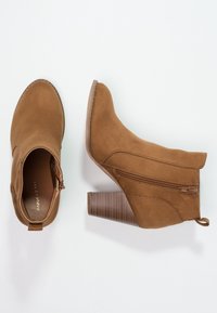 Botas de tobillo de ante marrón con un tacón bloque texturizado y cremallera lateral. El diseño cuenta con una puntera redondeada y una parte superior sin costuras.