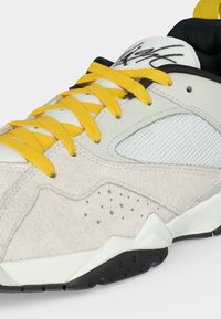 Λευκό και μπεζ αθλητικό παπούτσι με κίτρινα κορδόνια, μαύρη και λευκή σόλα, δίχτυ στο πλαϊνό πάνελ και μαύρο λογότυπο στη γλώσσα.