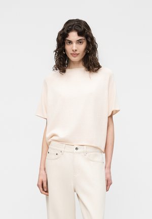 Kobieta o kręconych, ciemnych włosach, ubrana w luźny, kremowy top z krótkim rękawem oraz kremowe spodnie z wysokim stanem, na tle jednolitego, jasnego tła.