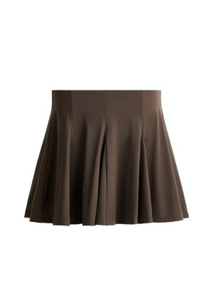 Femme debout portant une chemise à rayures marron à manches longues, une jupe plissée marron foncé, des chaussettes blanches, des baskets beiges et portant un sac à bandoulière blanc.