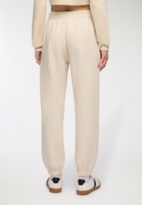 Pantaloni della tuta color crema con vita elasticizzata, polsini affusolati e una texture morbida, abbinati a sneakers bianche con dettagli neri.