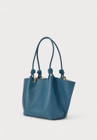 Kék bőrből készült bevásárlótáska két hosszú, csomózott fogóval és aranyszínű szerelékkel, egyszerű világos háttér előtt bemutatva.