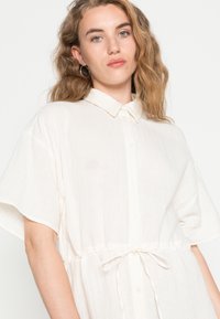 Vestido curto de mangas curtas em bege claro, com fecho de botões na frente, cintura elástica com cordão, feito de tecido texturizado. Sem padrões ou detalhes visíveis.