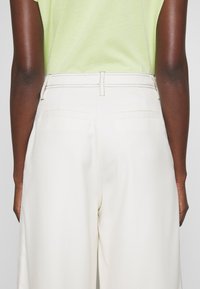 Vue arrière d'une personne portant un pantalon blanc taille haute avec des poches arrière cousues et un haut vert clair rentré à l'intérieur.