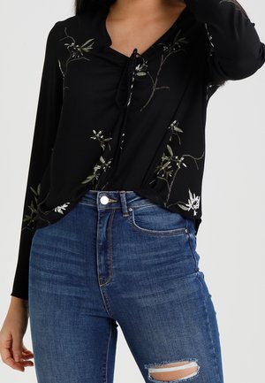 Kvinde iført en sort blomstret langærmet top stoppet ned i højtaljede blå jeans med en lille revne på det ene knæ.