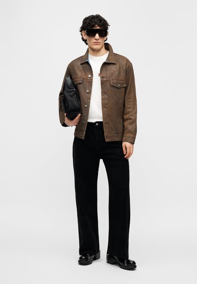 Καφέ δερμάτινο μπουφάν με κουμπιά, λευκό t-shirt, μαύρο παντελόνι κοτλέ και μαύρα γυαλιά ηλίου· κρατά μια μαύρη τσάντα.