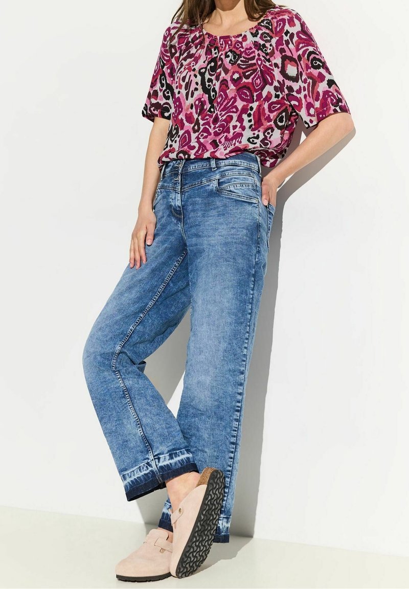 Femme portant un chemisier rose à manches courtes à motifs, un jean bleu taille haute et des chaussures beiges à enfiler, appuyée contre un mur blanc.