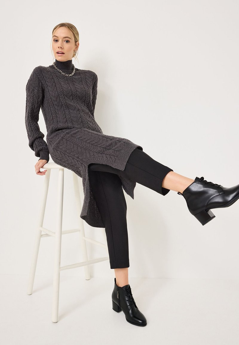 Abito di maglione grigio lavorato a maglia con motivo a trecce, spacco laterale, abbinato a pantaloni neri aderenti e stivaletti neri con tacco a blocco.
