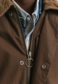 Bruine zip-up jas met een corduroy kraag, metalen rits en knoopaccenten, gelaagd over een blauw-wit gestreept T-shirt.