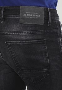 Mustat farkut, joissa on tyylikäs muotoilu, näkyvät saumat ja nahkapaikka, jossa lukee "JACK & JONES" vyötärön takana.