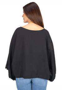 Schwarze, strukturierte Bluse mit weiten Ärmeln, offenem Rücken und Spitzenbesatz am Saum, kombiniert mit hellblauen Jeans.
