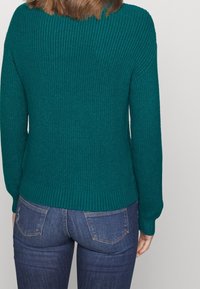 Teal stickad tröja med ribbad textur, rund halsringning och långa ärmar, kombinerad med mörkblå tajta jeans. Tröjans nederkant är ribbad.